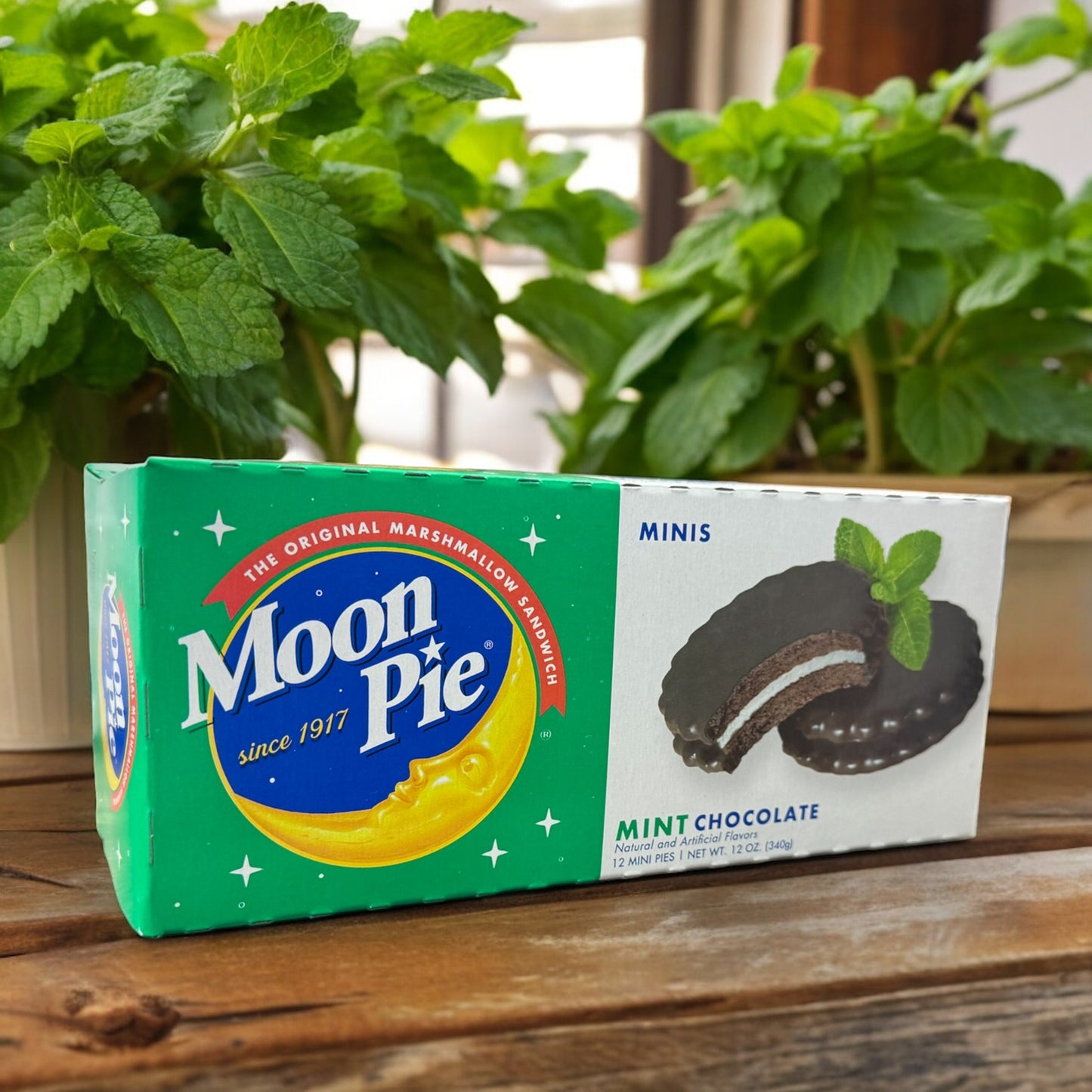 A box of Mini MoonPie mint chocolate cookies placed on a wooden surface with mint plants in the background.