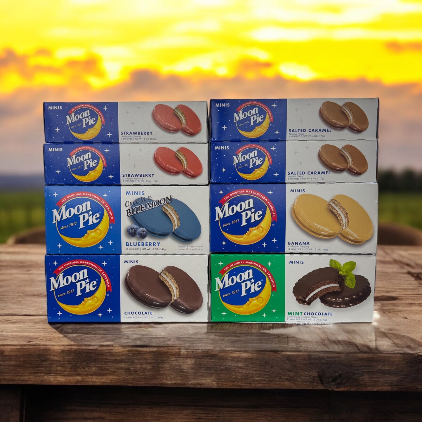 A collection of MoonPie mini snack cakes in various flavors, including chocolate, mint chocolate, blueberry, banana, salted caramel, and strawberry, displayed against a sunset background.